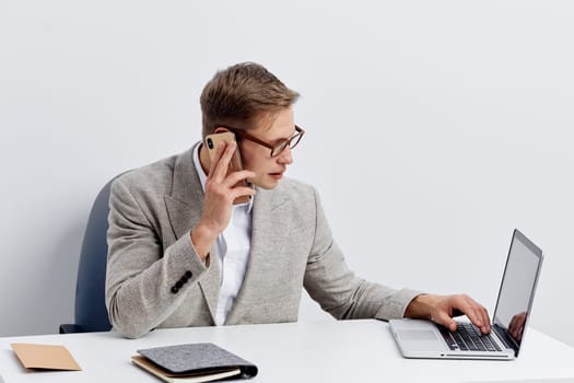 Middle-aged man dressed in business casual attire engaged in conversation on a smartphone while working on a laptop at a clean office desk, with a notepad and notebook, against a plain white background. Concept: professional communication, remote work.