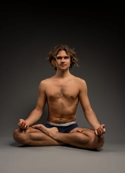 Full body shirtless young man in boxer briefs sitting in Lotus pose and looking at camera during meditation session against gray background