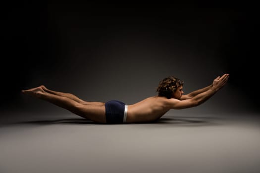 Side view of flexible young male in boxer briefs closing eyes and stretching during yoga session against dark gray background