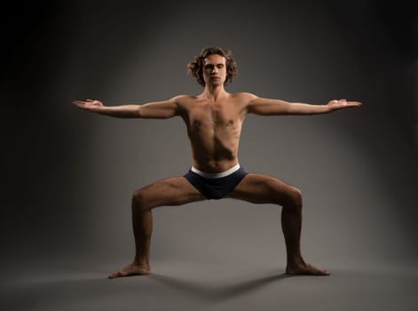 Side view of muscular male athlete in sportswear standing in warrior pose while doing yoga on gray background