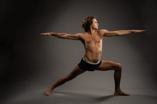 Full body barefoot young male with naked torso spreading arms and doing Virabhadrasana B pose during yoga session against gray background