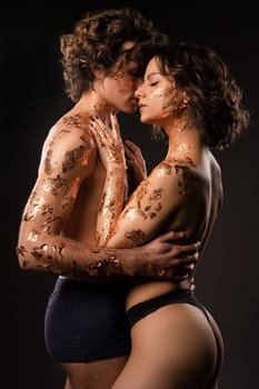 Side view of sensual topless couple in golden foils on bodies embracing gently while standing against dark