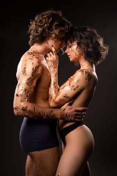 Side view of sensual loving couple in underwear with wavy dark hair and golden foils on bodies embracing gently while standing against black background