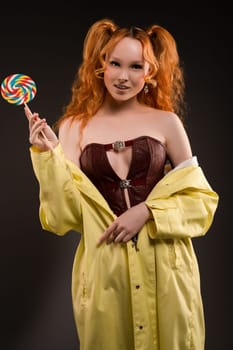 Positive sexy young female model with funny red curly pigtails in leather top and coat smiling while holding colorful spiral lollipop