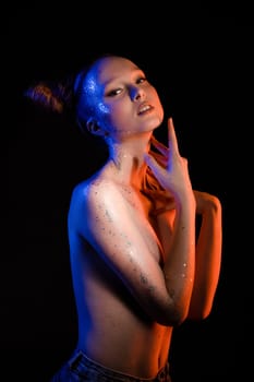 Side view of self assured young topless female model with creative hairstyle and glitter on body covering breast and looking at camera against black background