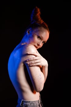Side view of self assured young topless female model with creative hairstyle and glitter on body covering breast and looking at camera against black background