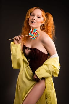 Positive sexy young female model with funny red curly pigtails in leather top and coat smiling while holding colorful spiral lollipop