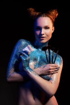 Self assured young female model with red hair topless painted with blue shiny glitter holding various makeup brushes and looking at camera