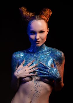 Sensual young female model with red hair covering breast with hands in blue bodyart in dark studio