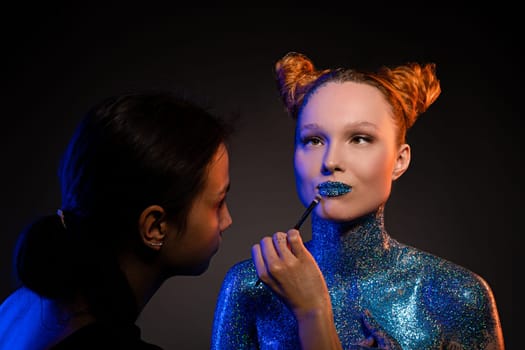 Side view of concentrated young female visagiste applying glitter on lips and naked body of confident model against black background in studio