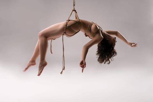 Full body of sensual naked female model tied up with rope against white background - bdsm shibari session