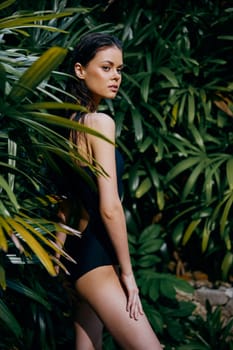 Attractive young woman in black swimsuit standing against lush green foliage background at tropical resort