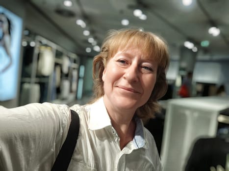 A middle-aged woman smiles happily while taking a selfie in a lively shopping area, reflecting her joyful experience exploring the location.