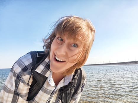 Middle-aged blonde woman smiles brightly while taking a selfie by the water on her travel excursion enjoying the sunny day.