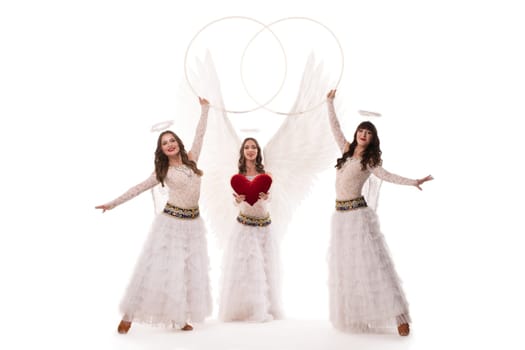 Full body of female dancer in stylish angel outfits with wings and halos looking at camera with hoops and heart against white background
