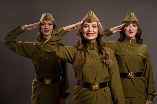 Positive women wearing military outfits looking at camera while making salute gestures with hands over temples against gray background in studio