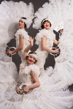 Beautiful female dancers in stylish burlesque costumes with headbands looking at camera with white feather fans in light modern studio