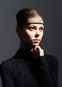 A contemplative young woman wearing a stylish black outfit and unique mesh headpiece, captured in soft dramatic lighting against a dark background, radiating beauty.