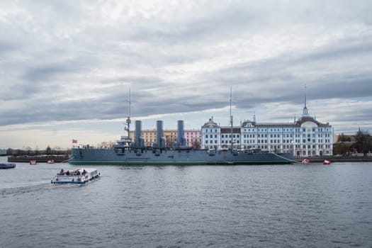 Saint Petersburg, Russia - 02.05.25. Iconic cruiser Aurora in front of the Nakhimov Naval School on a cloudy day. A small boat passes by, adding a dynamic element to the scene.
