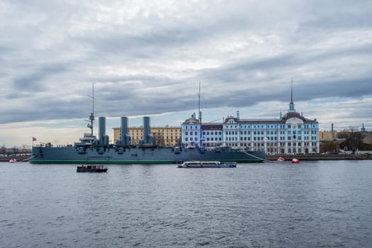 Saint Petersburg, Russia - 02.05.25. Iconic cruiser Aurora in front of the Nakhimov Naval School on a cloudy day. A small boat passes by, adding a dynamic element to the scene.