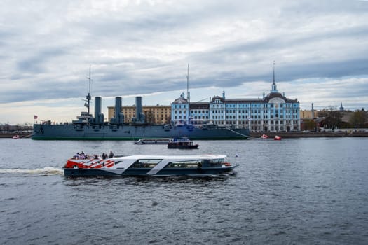 Saint Petersburg, Russia - 02.05.25. Iconic cruiser Aurora in front of the Nakhimov Naval School on a cloudy day. A small boat passes by, adding a dynamic element to the scene.