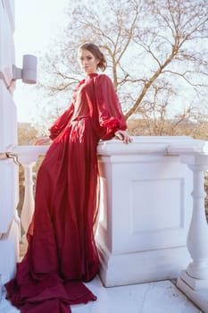 attractive girl in long red silk dress standing on balcony. sun rays falling on girl with beautiful makeup and hairstyle