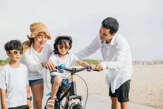 A perfect summer day unfolds as happy parents teach their children to ride bicycles on a sandy beach near sea. Smiles safety helmets and joy of family bonding create a cheerful tourism day concept.