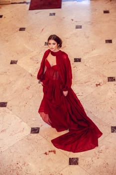 young and beautiful girl in a long evening red dress poses in the interior of the hotel. black dress with a corset and white sandals. she has an hairstyle and makeup