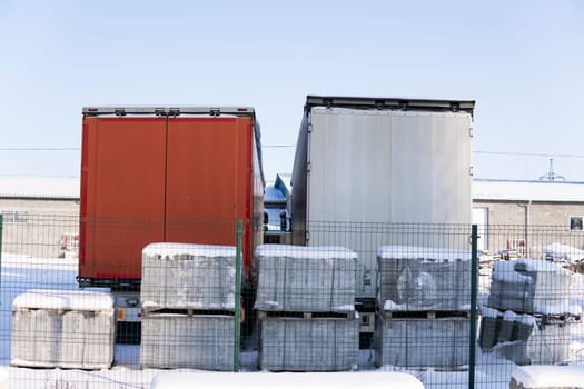 Rear view of red and white truck trailers parked near warehouse under bright sky, surrounded by snow-covered pallets and industrial fencing in cold weather.