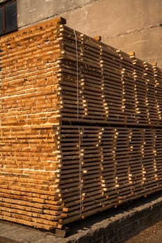 Large stack of dried wooden planks piled near a concrete wall. Industrial lumber ready for construction or carpentry, bathed in warm, golden afternoon light.