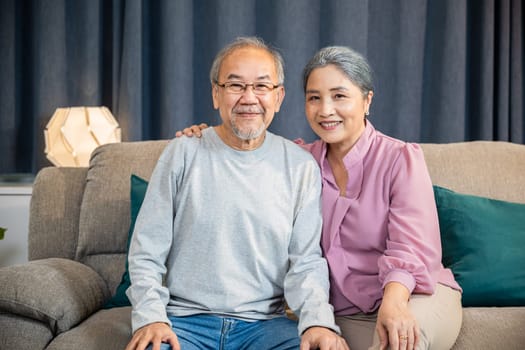 Portrait of Happy Asian senior couple greeting someone while sitting on a comfortable couch, old man and woman sit close together on a sofa in the house