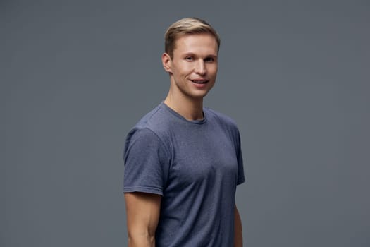 Happy young man with short blond hair dressed in a casual blue t-shirt, smiling confidently, posed against a neutral gray studio backdrop. Concept: positive attitude and relaxed style