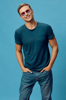 Cheerful woman with short blonde hair, dressed in a comfortable blue t-shirt and jeans, standing confidently with hands in pockets. Bright pink sunglasses enhance her joyful, lively appearance in a studio setting. Concept: positivity and casual fashion.