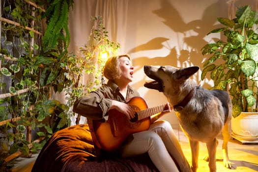 A woman sings joyfully to her dog while playing guitar in a warm, inviting room filled with plants and soft lighting.