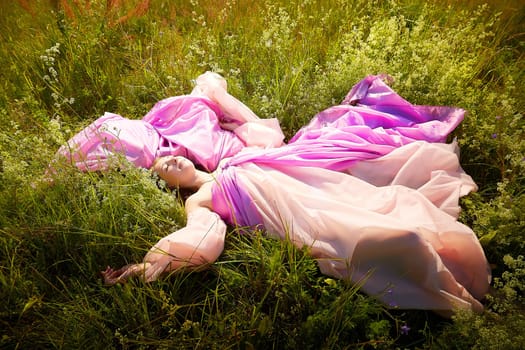 A woman lies on her back in a vibrant field, wearing a billowing pink gown that contrasts with the lush greenery and wildflowers around her.