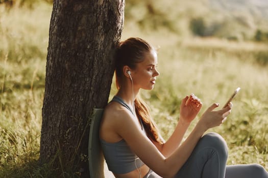 A young woman listens to music with earbuds in a fitness outfit, sitting against a tree in a lush green area, radiating happiness and tranquility in a bright, sunny atmosphere