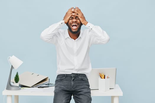 Happy man with dark skin and beard, wearing a white shirt and jeans, standing at office desk, covering face with hands, showing frustration or stress, in a clean workspace with pastel blue background. Concept: workplace emotion or work stress.