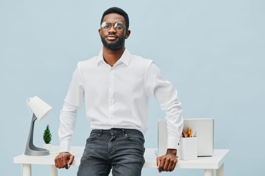 Stylish young man wearing a white shirt and black jeans stands confidently at a sleek desk with tech accessories and office supplies, set against a plain light blue background. Concept: professional workspace