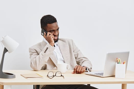 Concise image of a smiling young African American man in professional attire sitting at a desk with a laptop, holding a phone, surrounded by office supplies, expressing productivity and communication in a corporate setting. Concept: modern business communication