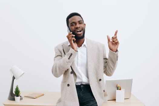 Confident smiling man engaging in a phone conversation while standing at a desk in a contemporary office environment with minimal decor, laptop, and stationery, illustrating effective communication, modern office, casual professional style, cheerful atmosphere, studio shot, concept: business communication.