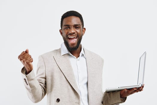 Happy man with dark skin wearing beige blazer and white shirt, holding a laptop and expressing excitement, standing against a plain white backdrop. Concept: success and enthusiasm.