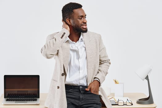 Young professional man with dark curly hair, wearing a beige blazer and white shirt, standing in a contemporary office environment and smiling. He appears relaxed and confident, with a laptop, glasses, and office supplies on the desk. Concept: workplace success.