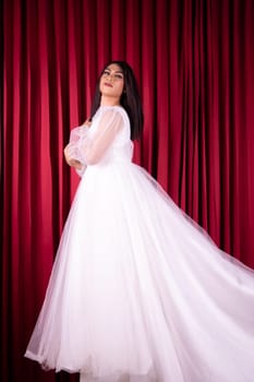 Beautiful Asian woman standing with a happy face in a white wedding dress in front red curtain inside the studio