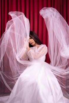 Elegance Asian woman wearing a wedding dress with flying fabric around her in front of the red curtain inside the studio