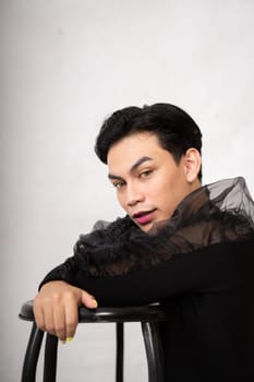 Pretty Asian man putting his hand and head on the chair while posing in a black dress inside the studio