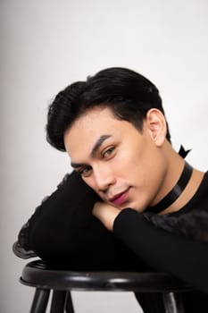 Pretty Asian man putting his hand and head on the chair while posing in a black dress inside the studio