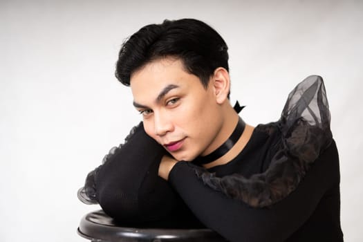 Pretty Asian man putting his hand and head on the chair while posing in a black dress inside the studio