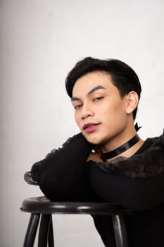 beautiful Asian man posing with the chair while wearing a black costume and makeup inside the white studio