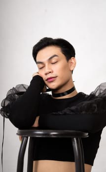 beautiful Asian man posing with the chair while wearing a black costume and makeup inside the white studio