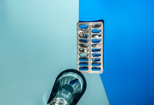 Medicine in blue capsules on a blue background. The background is light and dark blue. A glass of water and capsules in a blister. Space for text, top view. The concept of a medical pharmacy